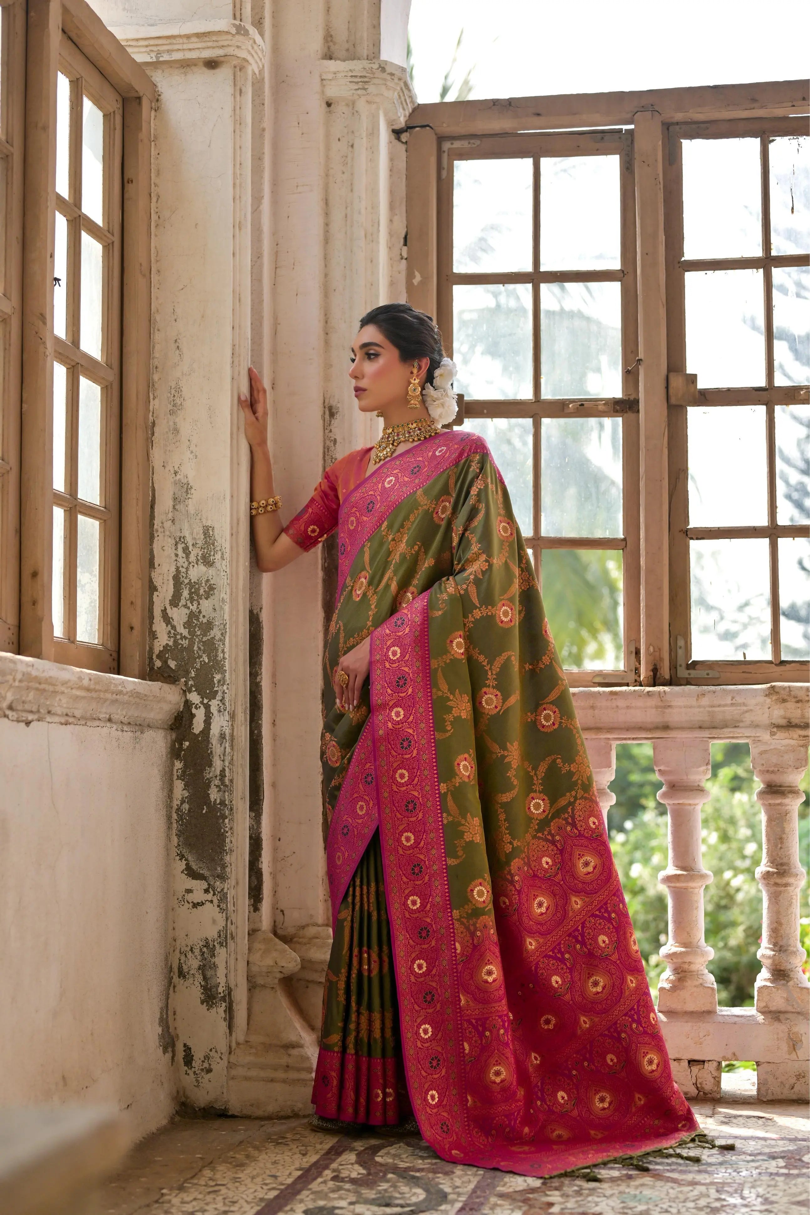 A woman models a striking Mehandi Green Banarasi Silk Saree with gold Zari weaving. The saree has a wide, ornate Fuchsia Pink border and pallu featuring dense traditional geometric Zari weaving. She wears a coral pink blouse and heavy gold Kundan jewelry.